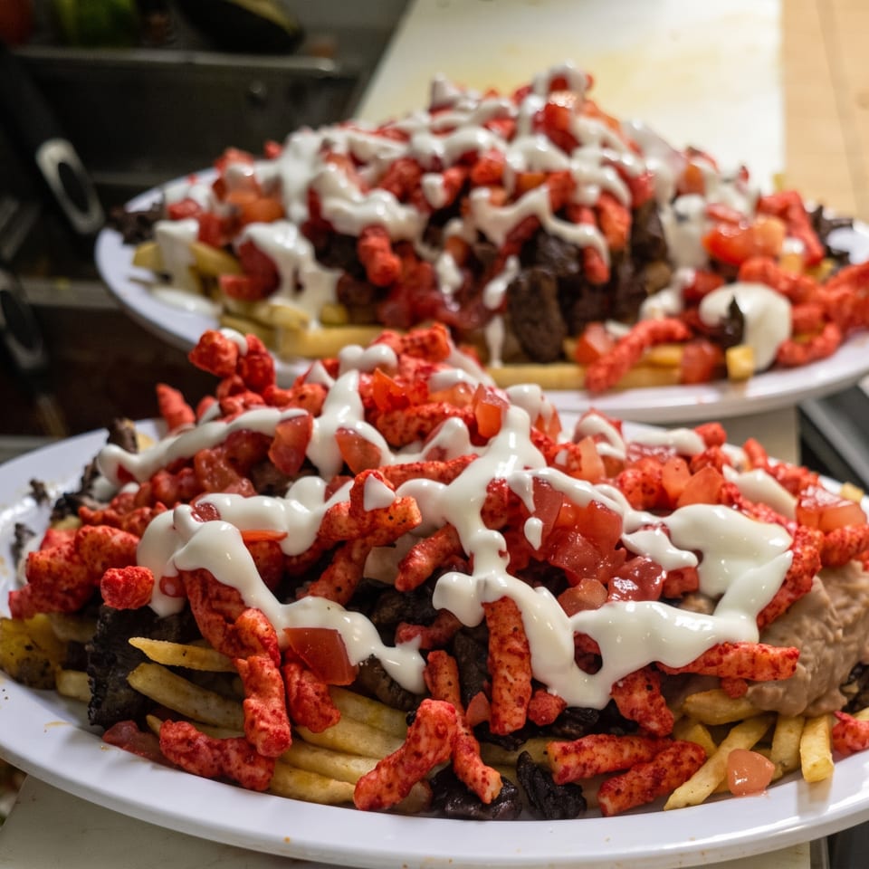 Two plates of loaded fries topped with hot Cheetos, diced tomatoes, and white sauce.