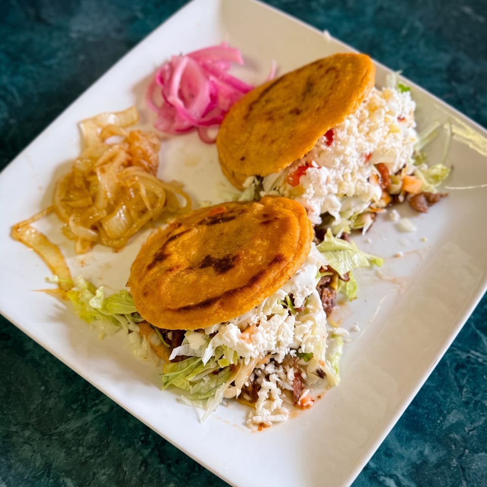 Two golden pupusas filled with lettuce, cheese, and meat, served with pickled onions.